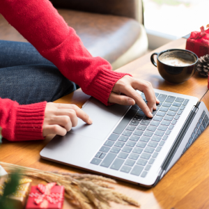 A woman dressed in red sweater typing on a lapptop near a cup of coffee and gifts. Overcome Holiday Impulse Purchases